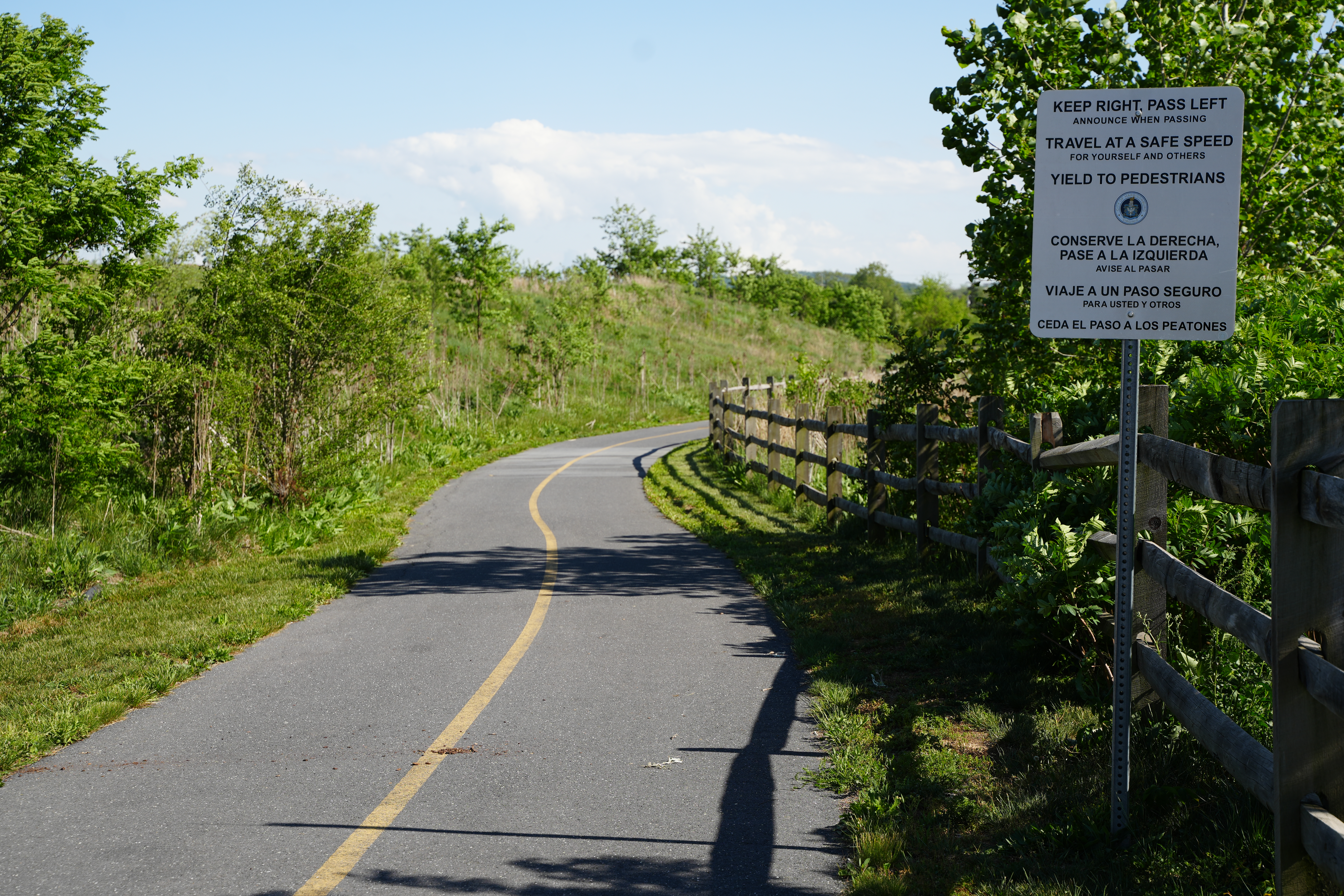 paved path with split rail fence on right side. Bushes and small trees line both sides of the trail.